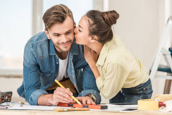 young woman kissing smiling man marking blueprint with level tool 
