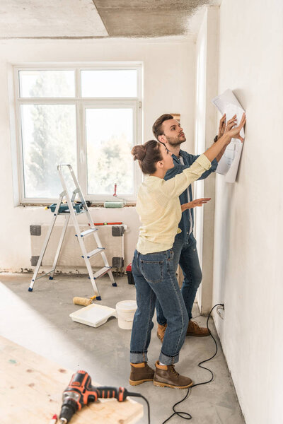 side view of young couple holding blueprint at wall during renovation