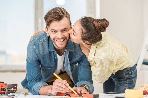 young woman kissing happy man marking blueprint with level tool 