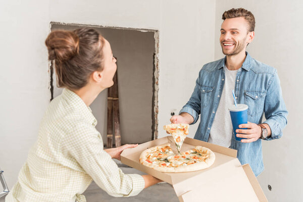 happy young couple holding box with pizza and paper cup with refreshing beverage in new apartment