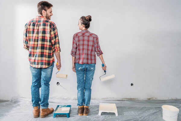 back view of young couple holding paint rollers and smiling each other during renovation