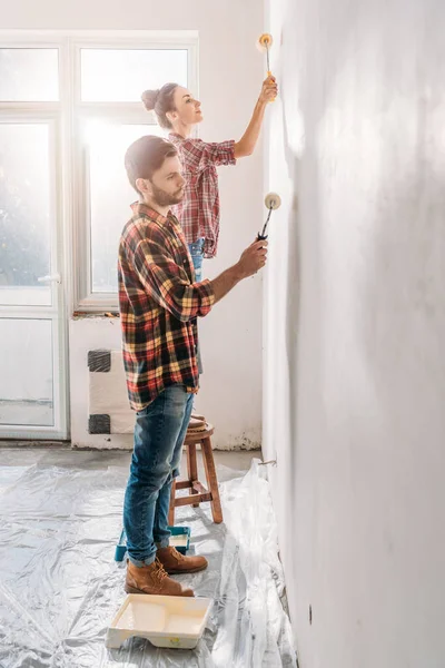 side view of young couple holding paint rollers and painting wall in new apartment