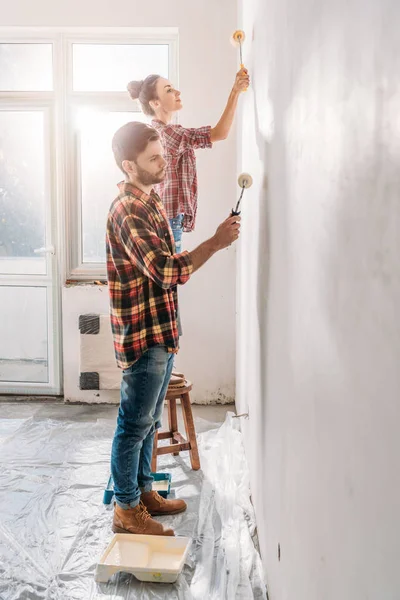 在新公寓举行油漆辊和油漆墙的年轻夫妇的侧视图 — 图库照片