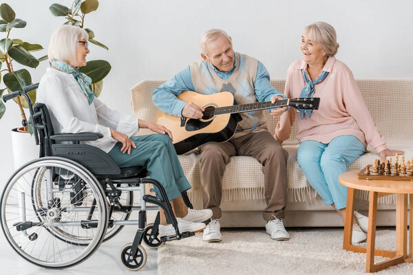 старший мужчина сидит на диване и играет на акустической гитаре для женщин
