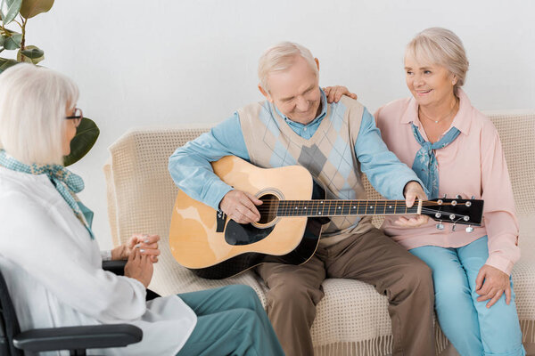 senior man sitting on sofa and playing acoustic guitar for women