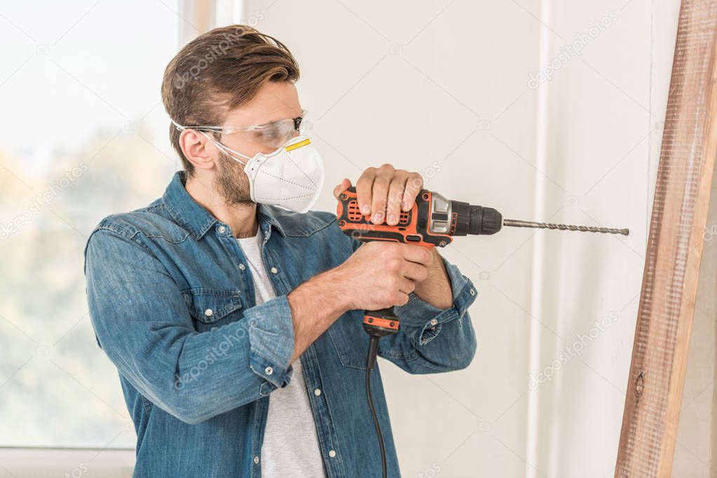 young man in protective mask and goggles using electric drill during house repair