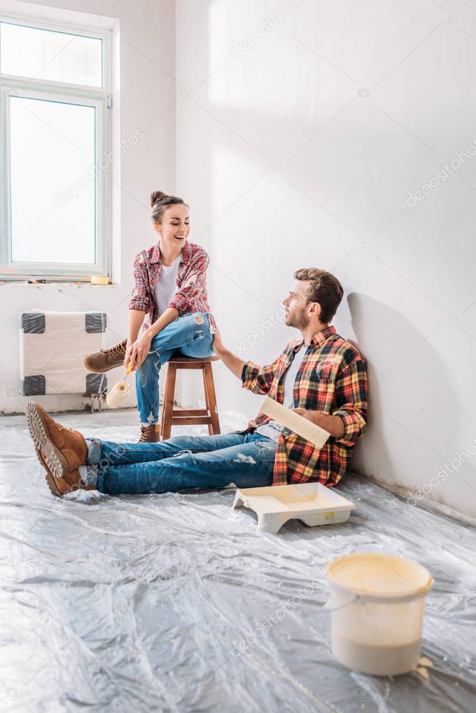 smiling young couple holding paint rollers and talking in new apartment