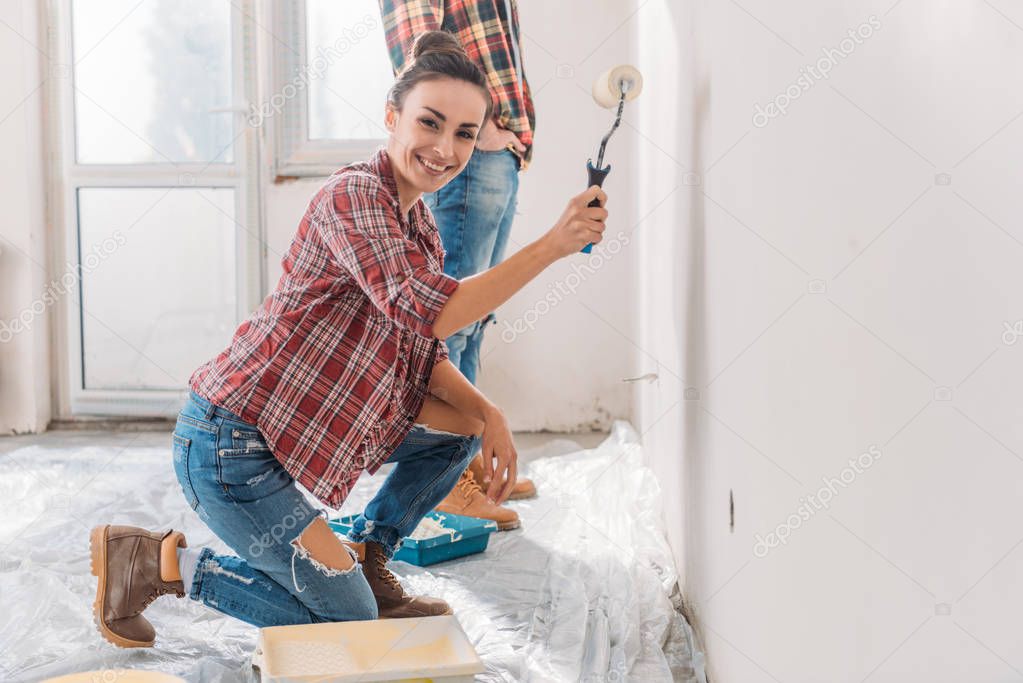 happy young woman painting wall and smiling at camera in new apartment