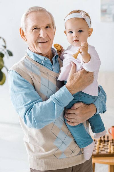 senior grandfather holding toddler granddaughter with cookie and chess figure in hands