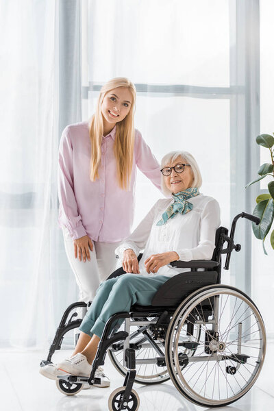 young woman standing near senior woman in wheelchair and looking at camera