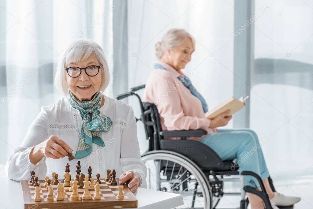 senior woman playing chess at table while another sitting in wheelchair and reading book