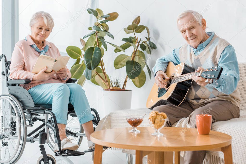 senior woman sitting in wheelchair and reading book while senior man playing acoustic guitar