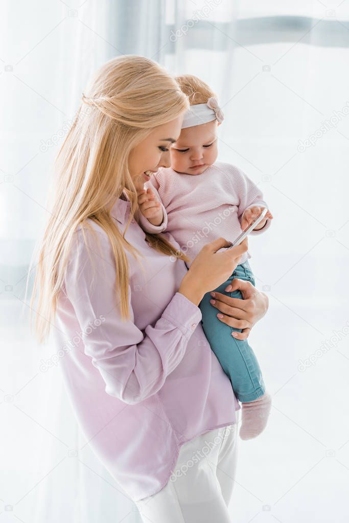 happy mother holding toddler and using smartphone