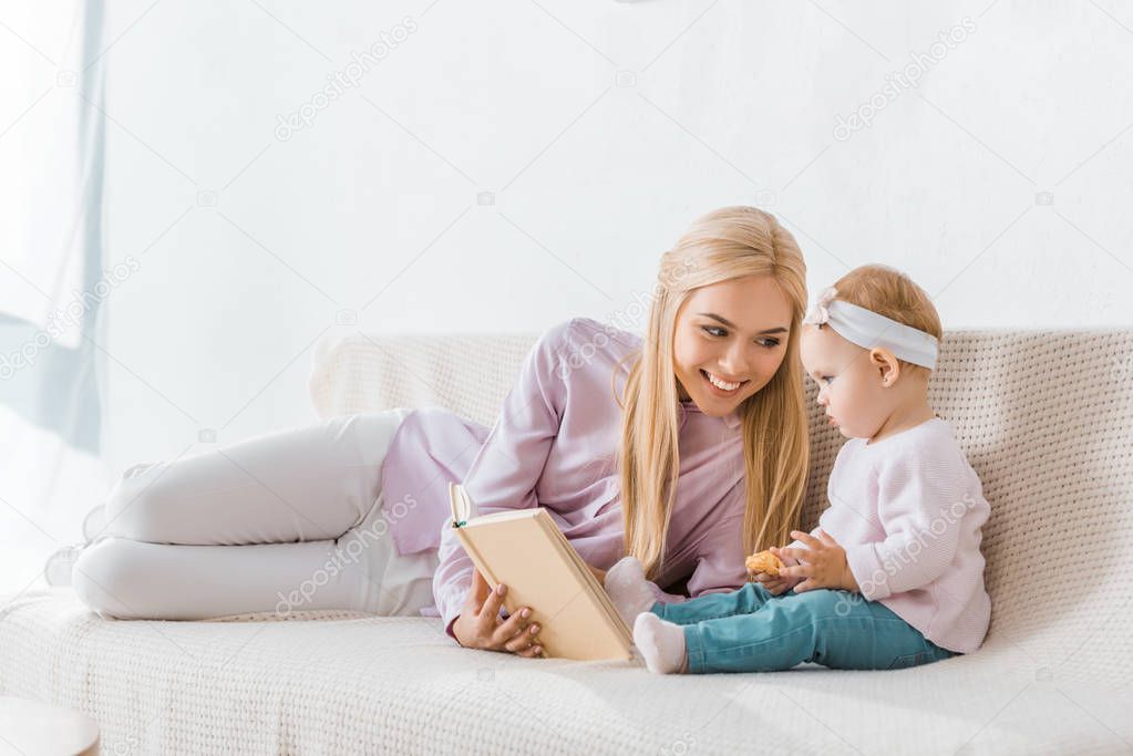 young smiling mother reading book to small daughter while toddler holding cookie