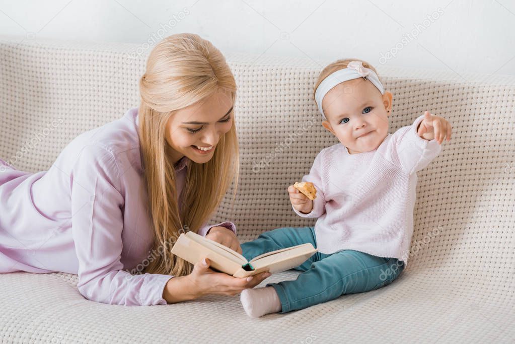 young happy mother laying on sofa and reading book to small daughter while toddler holding cookie