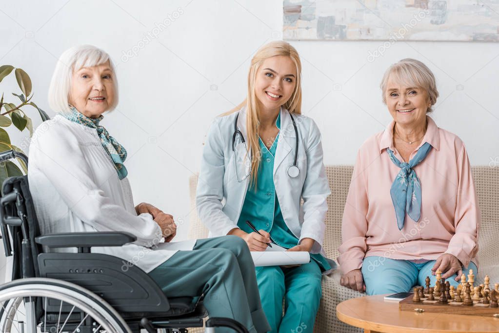 young female nurse sitting on sofa and writing prescription to senior women