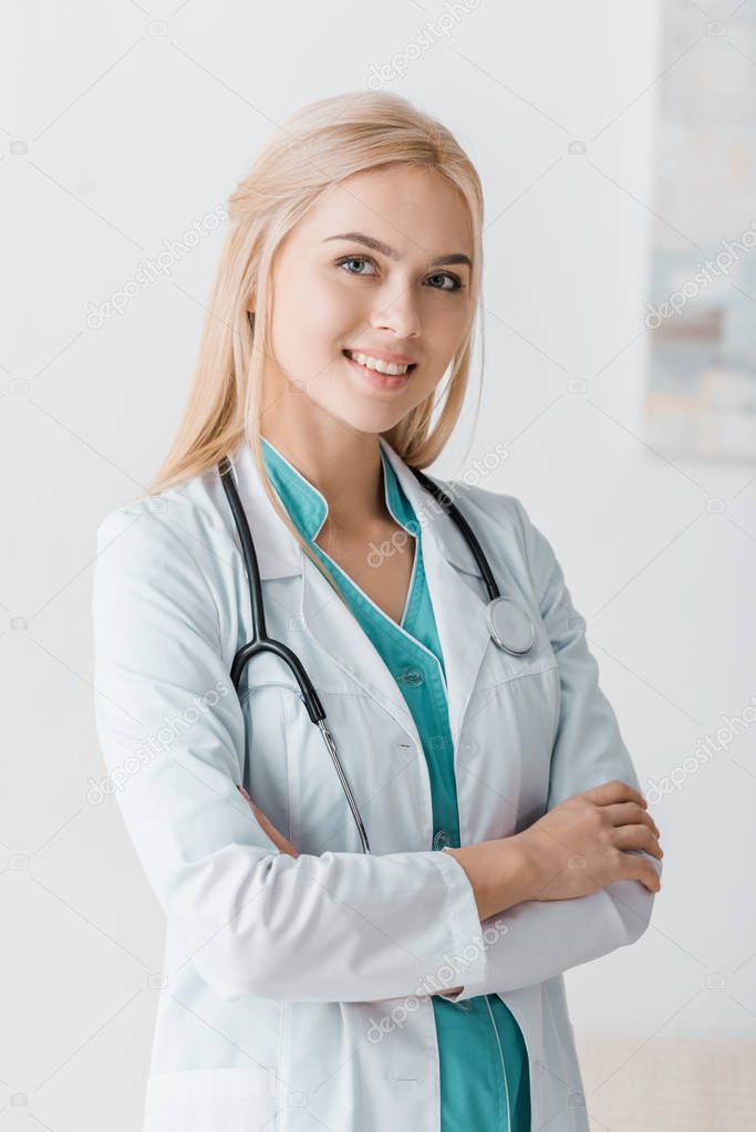 smiling young female doctor with stethoscope standing with arms crossed in clinic 