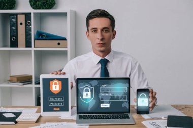 portrait of businessman showing laptop, tablet and smartphone with cyber security signs on screens at workplace in office clipart