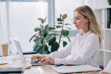 side view of businesswoman in eyeglasses working on laptop at workplace in office clipart