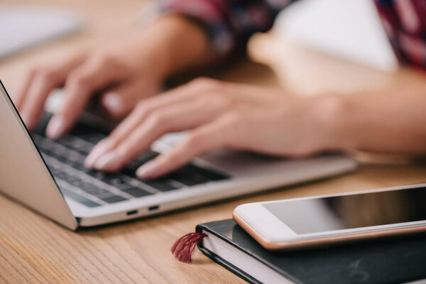 partial view of woman typing on laptop at tabletop with smartphone and notebook