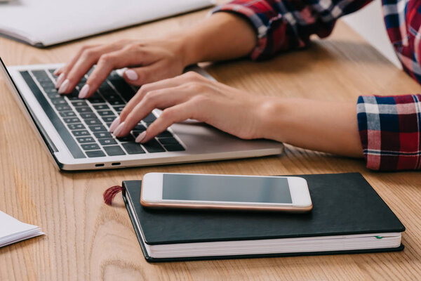 partial view of woman typing on laptop at tabletop with smartphone and notebook