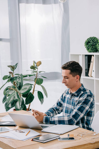 side view of businessman using smartphone at workplace with laptop in office and notebook