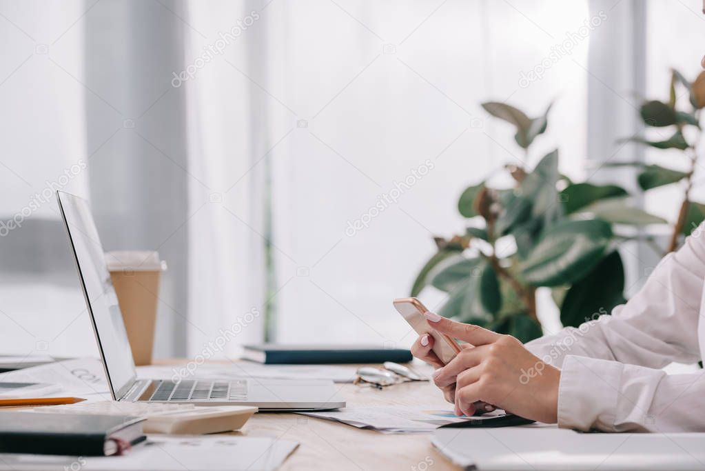 partial view of businesswoman with smartphone at workplace with documents and laptop in office
