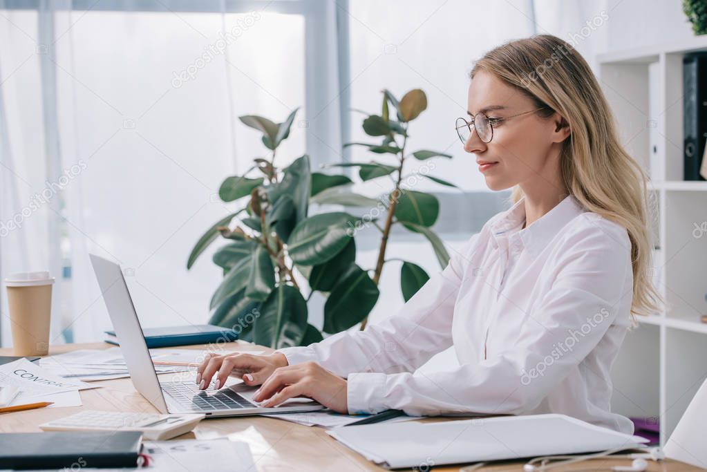side view of businesswoman in eyeglasses working on laptop at workplace in office