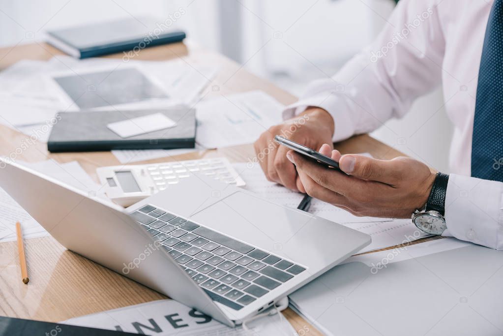 partial view of businessman using smartphone at workplace with papers and laptop