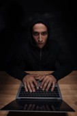 portrait of hacker in black hoodie looking at camera while using laptop, cuber security concept