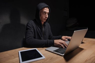 hacker in black hoodie using laptops at tabletop with tablet in dark room, cyber security concept clipart