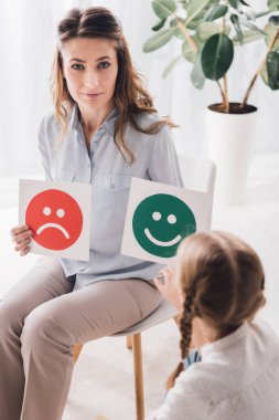 adult psychologist showing happy and sad emotion faces cards to child and looking at camera clipart