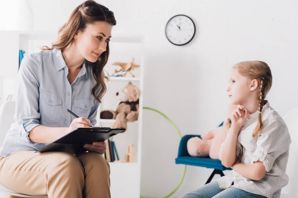 adult psychologist with clipboard sitting in front of little child in office