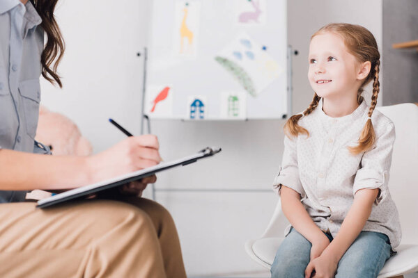 cropped shot of psychologist with clipboard sitting in front of child in office