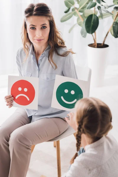 adult psychologist showing happy and sad emotion faces cards to child and looking at camera