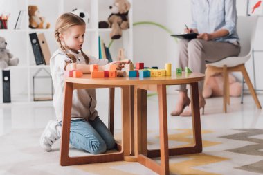 concentrated little child playing with blocks while psychologist sitting blurred on background clipart