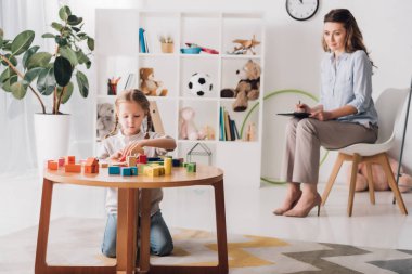 little child playing with blocks while psychologist sitting blurred on background clipart