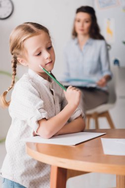 thoughtful little child drawing while psychologist with clipboard sitting blurred on background clipart