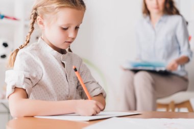 concentrated little child drawing while psychologist with clipboard sitting blurred on background clipart