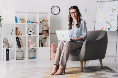 Çocuk psikologu ise office sandalyede oturan ve kameraya bakarak laptop ile çalışma