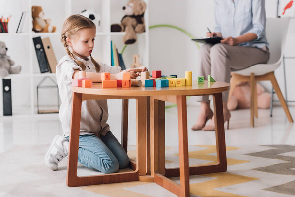 concentrated little child playing with blocks while psychologist sitting blurred on background