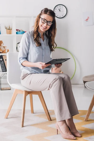 Lachende Kinderpsycholoog Met Klembord Zit Stoel Office Camera Kijken — Stockfoto