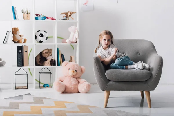 孤独な子供のおもちゃの棚の前で椅子に座っているとカメラ目線 — ストック写真