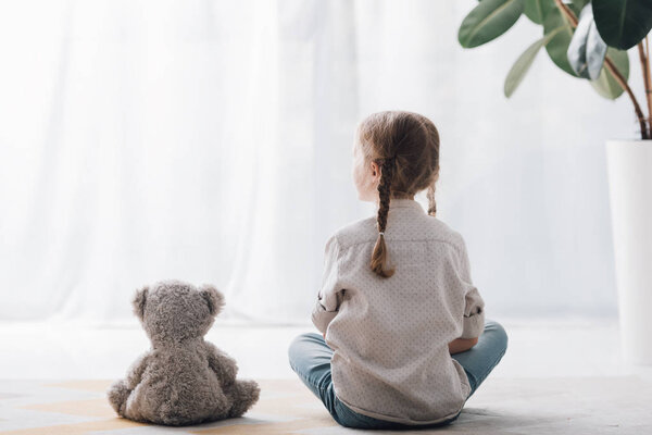 rear view of little child sitting on floor with teddy bear and looking away