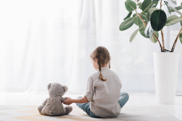 rear view of little child sitting on floor in front of curtains with teddy bear