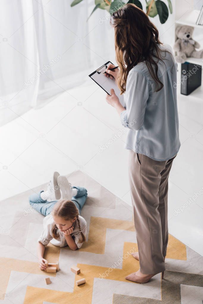 high angle view of psychologist with clipboard standing near child while she lying on floor and playing with wooden blocks