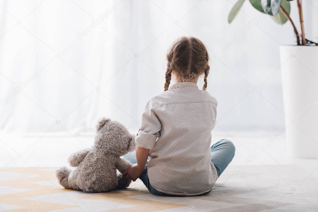 rear view of little child sitting on floor with her teddy bear toy