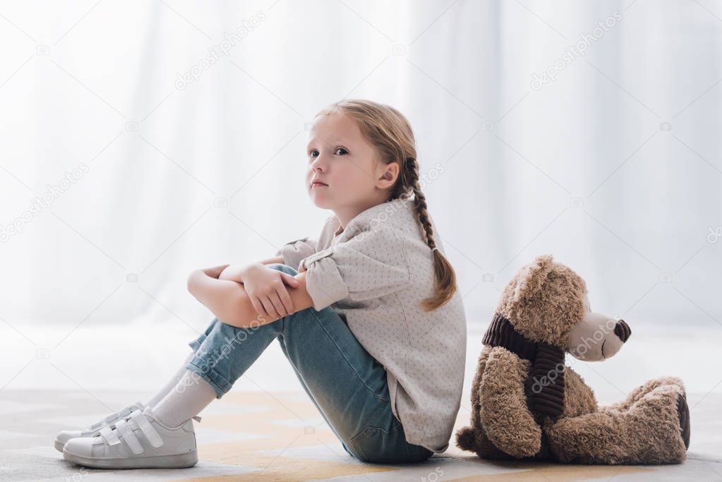side view of depressed little child sitting on floor back to back with teddy bear