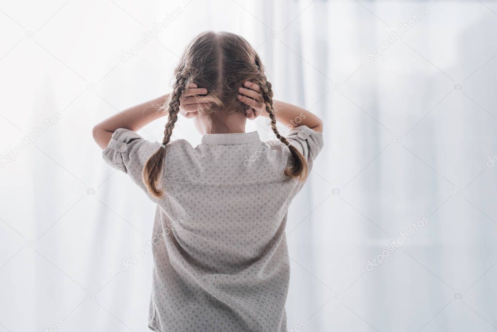 rear view of little child covering ears with hands while standing in front of curtains