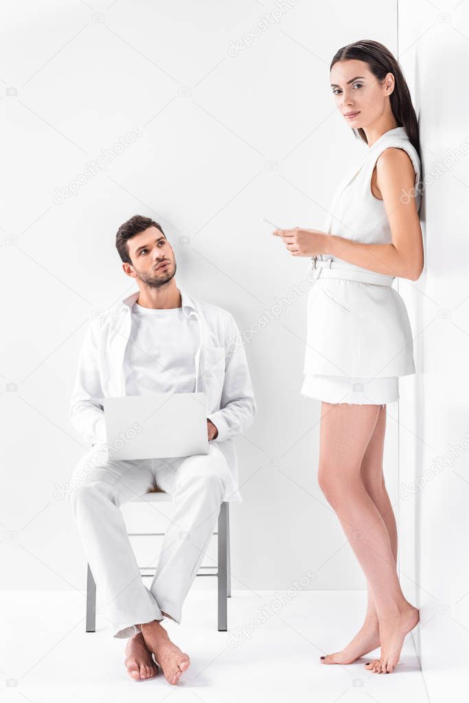 adult man in total white sitting on chair and typing on laptop while attractive woman using smartphone